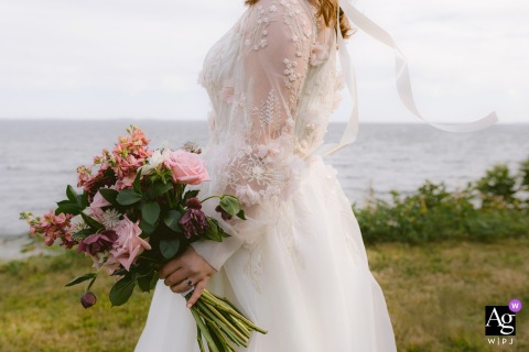   The bride poses at her Camden, Maine ceremony location, holding her bouquet as the ties from her dress flutter in the breeze behind her, adding movement to the scenic detail.