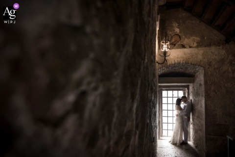 Silhouetted Near Barred Window Inside Malcesine Castle On Lake Garda, The Couple Shares A Tender Embrace In Shadows Inside Malcesine Castle on Lake Garda, Italy, the couple is silhouetted in a tender embrace near a barred window, soft shadows from the ancient stone walls creating a romantic scene.