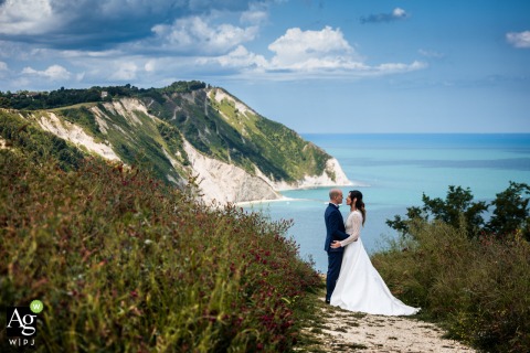 A Couple Portrait Pose With Monte Conero Views At Portonovo Majestic Seaside Backdrop A couple poses for a portrait with Monte Conero’s breathtaking landscape as their backdrop at Portonovo, the sweeping views lending natural majesty to the seaside image.