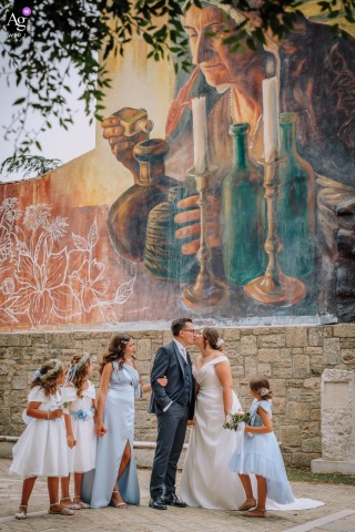 Beneath A Monumental Urban Mural In Apulia, The Couple Poses Together With Children As Artwork Shapes Their Meaningful Scene Setter Beneath a monumental mural at their ceremony location in Apulia, the couple stands together, the striking urban artwork weaving personal and collective stories into their wedding day narrative.