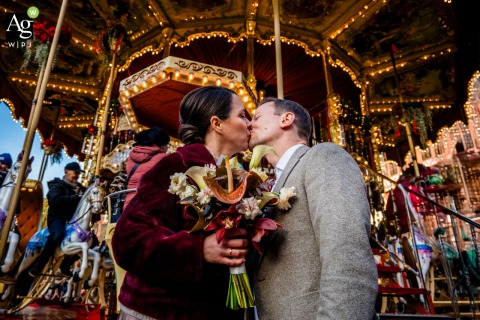 Couple Kisses In Front Of Carousel At Frankfurt Christmas Market Roemer Germany Under twinkling lights at the Frankfurt Christmas Market Roemer, a couple stands and kisses before a carousel, sharing a festive and whimsical winter portrait session.