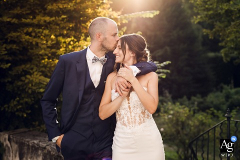 Bride Holds Her Partner’s Hands As He Kisses Her Forehead At Sunset, Golden Light Enveloping Their Portrait At the reception venue, the newlyweds stand together at sunset, the bride holding her partner’s hands as he gently kisses her forehead in golden evening light.