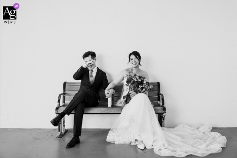 The Couple Shares Laughter On A Bench At The Groom’s Home In Singapore In A Lighthearted Portrait At the groom's home in Singapore, the couple sits together on a bench, sharing a lighthearted portrait as the groom laughs with visible joy.