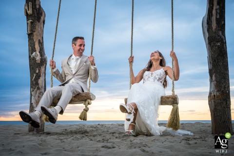 Bride And Groom Share Laughter Swaying On Beach Swing At Makani Beach Ter Heide At Makani Beach in Ter Heide, the bride and groom share laughter and playfulness as they sway together on the tall beach swing, savoring lighthearted fun at their wedding.