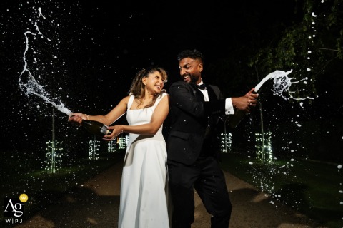 Bubbles And Champagne Spray Envelop The Couple At South Farm, Adding Playful Joy To Their Night Portrait. Bubbles and joy surround the couple at South Farm as they share a champagne spray portrait, the rural backdrop adding a playful spirit to the scene.