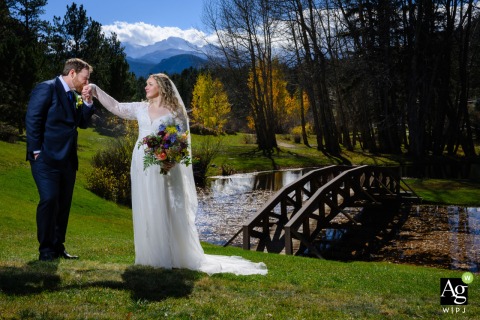 Groom Kisses Bride’s Hand Beside Black Canyon Inn Pond In Estes Park Colorado Beside a tranquil pond at Black Canyon Inn in Estes Park, Colorado, the groom gently kisses the bride’s hand as towering evergreens and mountain scenery create a romantic backdrop.