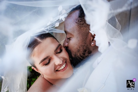 Couple Stands Beneath Bride’s Veil In Soft Intimate Portrait At Urla Sahne Beneath the bride’s veil at Urla Sahne in Urla, Izmir, Turkey, the couple stands close, the sheer fabric diffusing light for a soft and intimate wedding portrait.