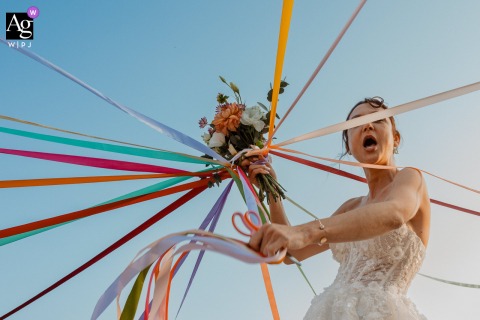   At Château du Parc Saint Lambert la Potherie, France, the bride holds her bouquet while cutting the colorful ribbons of her bridesmaids, preparing for the traditional bouquet round in a festive scene.