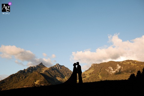 Bride And Groom Silhouetted Against Majestic Mountain Backdrop In Fribourg In Fribourg, the bride and groom appear as silhouettes at the reception venue, with a breathtaking mountain backdrop, bringing a dramatic sense of place and scale to their portrait.
