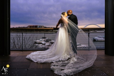Riverside Kiss At District Winery DC With Drifting Veil And Expansive Sky At District Winery in DC, the couple’s riverside kiss is set against a drifting bridal veil and an expansive sky, bringing drama and promise to their wedding portrait.