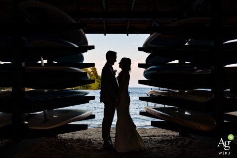 Bride and Groom Silhouetted in a Boathouse Between Paddleboards at Camp Mataponi, Maine, on Their Adventurous Wedding Day The bride and groom are silhouetted between paddleboards at Camp Mataponi in Maine, their figures outlined against the light on their adventurous wedding day.