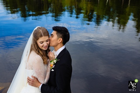 Groom Kisses Bride with Natural Beauty and Water Reflections at New England Outdoor Center, Millinocket, Maine A groom gently kisses his bride during their wedding portraits at New England Outdoor Center in Millinocket, Maine, with natural beauty and love shining through the water scene.