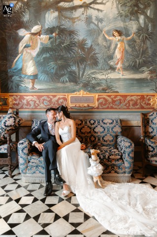 Little Dog Watches Newlyweds Beneath Ancient Painting At Castello Di Torre Alfina On a classic sofa beneath a large, ancient painting inside Castello di Torre Alfina in Viterbo, the newlyweds draw near, while a little dog curiously looks on at their intimate session.