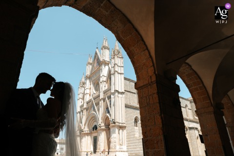 Silhouetted Spouses Draw Near Under Graceful Arches Before Orvieto Cathedral's Iconic Façade Under the graceful arches of the portico in front of Orvieto Cathedral, the two spouses are silhouetted as they draw near to each other, with Orvieto’s iconic façade in the distance.