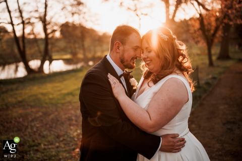 Winter Sunsets Bathes Bride and Groom in Gentle Light by Rushton Hall Hotel and Spa, England, UK Beneath tall, wintry trees just beyond Rushton Hall Hotel and Spa, England, UK, the bride and groom stand close together as the sunset bathes the woodland in gentle light.