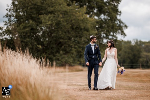 Bride And Groom Share Connective Glance In Lush Gardens Of Burningfold Barn In the lush gardens of Burningfold Barn, England, the bride and groom walk hand in hand, sharing a connective glance as greenery surrounds their wedding portrait.