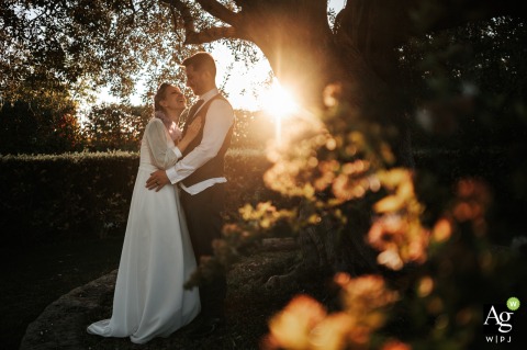Bride Laughs Joyfully in Groom’s Arms Beneath Sprawling Tree at Borgo Colle Mentuccia in Rome, Italy As golden sunset light filters through the branches at Borgo Colle Mentuccia in Rome, the bride laughs joyfully in the groom’s arms beneath a sprawling tree.