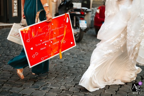 Red Welcome Sign And Flowing Dress On The Historic Streets Of Rome Walking the historic streets of Rome between church and restaurant, a friend of the bride holds a red wedding welcome sign beside the flowing fabric of the bride’s dress, marking the day’s joyful proceedings.