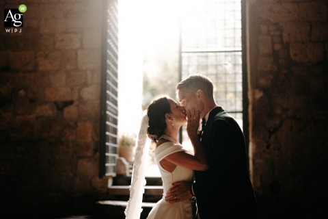 Newlyweds Kiss With Radiant Backlight Inside Serene San Giusto Abbey in Tuscania, Lazio, Italy In the serene stillness of San Giusto Abbey in Tuscania, Lazio, the newlyweds share a kiss inside the empty church, radiant backlight streaming through the open doors behind them.