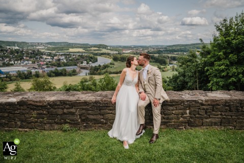Couple Shares Affectionate Kiss atop Ancient Wall at Burgruine Volmarstein, Germany, Surrounded by Historic Scenery Seated atop the ancient wall at Burgruine Volmarstein in Germany, a wedding couple shares an affectionate kiss, surrounded by the historic castle’s weathered stones and sweeping hillside scenery.