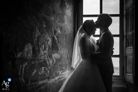 Castle Windows Illuminate The Couple At Château De Bagnols In France's Beaujolais Region At Château de Bagnols in France's Beaujolais region, a backlit portrait of the bride and groom uses soft light streaming through the castle windows to reveal the rich texture of the historic stone walls while framing the couple in a timeless, romantic sce
