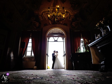 Historic Doorway Frames The Couple's Quiet Embrace At Villa Canton In Italy At Villa Canton in Italy, the bride and groom share a quiet portrait framed by the grand doorway of the historic villa, where elegant architecture, rich interior details, and dramatic backlighting draw the eye to their intimate embrace.