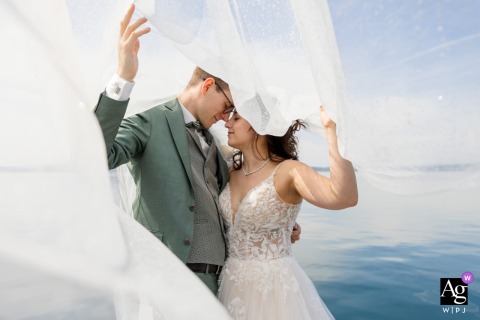 At Lake Zug, Switzerland, the bride and groom stand at the water's edge as the bride's veil lifts gracefully above them, creating a striking portrait that blends movement, elegance, and the tranquil lakeside setting.