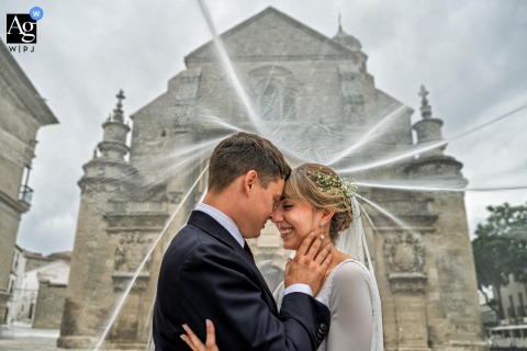 Flowing Veil Frames The Couple Before Historic Architecture In Úbeda, Jaén, Spain In Úbeda, Jaén, Spain, the bride and groom stand before a majestic old stone building as her veil stretches around them, softly separating the couple from the historic backdrop.