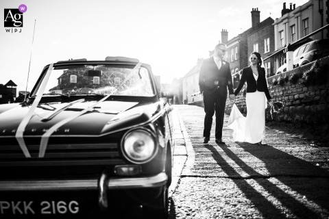 In Twickenham, London, England, the bride and groom walk together in soft backlight, while a car on one side of the frame adds an urban element to the elegant wedding-day portrait.