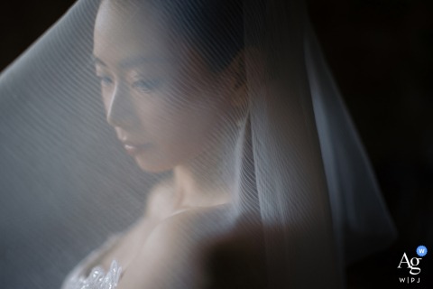 At the bride’s home in Maoming, Guangdong, China, she stands quietly by a window with her face softly visible through her veil, patiently awaiting the groom’s arrival before the wedding ceremony.
