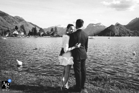 At Lake Annecy in Haute-Savoie, France, following their hotel ceremony, the bride and groom stand at the water’s edge surrounded by alpine scenery as the bride glances back toward the camera during their portrait session.