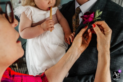 At an Airbnb in Lincoln, United Kingdom, buttonholes are carefully pinned onto the wedding party while a young guest casually enjoys breadsticks in the background, adding a humorous touch to the preparations.