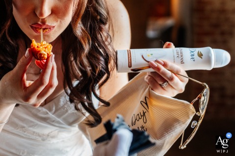 At Woolas Barn in Yorkshire, United Kingdom, the bride grabs a quick bite of arancini while holding a can of deodorant, creating a humorous and relatable moment during the wedding day preparations.