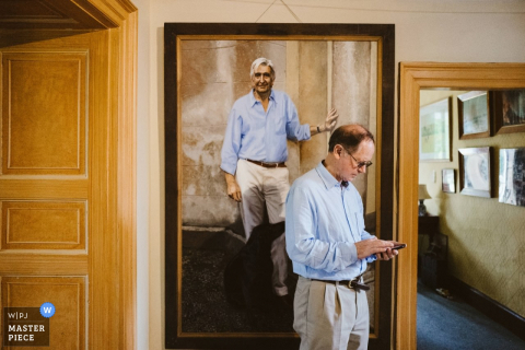 Photo of a man checking his phone in front of a painting of a man dressed identically to him by a Greater Manchester wedding reportage photographer.