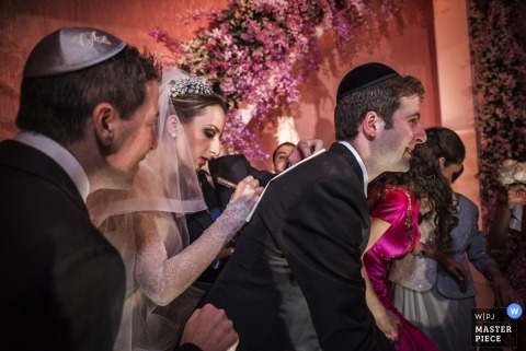 An image of a traditional bride using her smiling groom's back to write a quick note with an arch of wedding flowers in the background