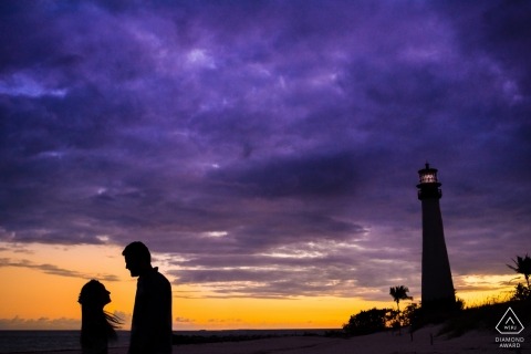 A vibrant purple and yellow sky create a dramatic backdrop for this lighthouse and silhouetted couple for their engagement portrait