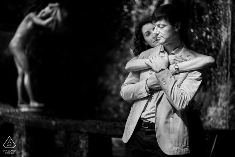 The engaged couple is posing with a statue watching them, with their arms all wrapped up in the photo an engaged couple and a statue watching them, Italy, Vittoriale degli Italiani, Garda Lake.