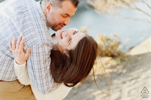 A young NY woman joyfully smiles as a young man dips her down in an embrace. Engagement Portrait by a New York Lake at Sunset 