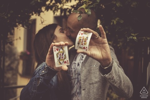 A couple holds up a Queen of Hearts and King of Hearts as they kiss beneath a tree in Bolano during their pre-wedding session by a Liguria photographer.
