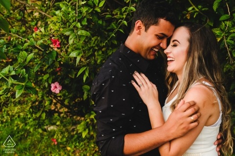 A couple laughs as they stand together in Ouro Preto by a green bush with pink flowers in this engagement session by a Minas Gerais, Brazil photographer.