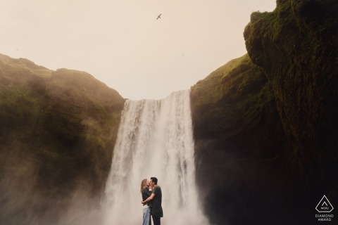 Engaged couple kissing with large Iceland waterfall in the background and a bird in the air 