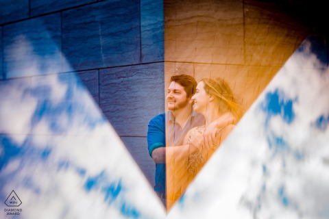 DC bride and groom to be, modeling for a pre-wedding picture at the National Gallery of Art in the East Building as the couple embrace, looking left, on the corner of the building, framed in a triangular reflection