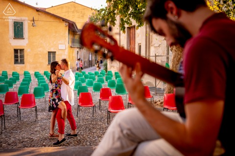 An after the wedding proposal engagement picture session at Iseo Lake in Italy as a Couple kisses near a man playing a guitar