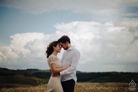 An intimate portrait of a couple as they embrace in a grassy field, with majestic clouds serving as a dramatic backdrop, during their engagement session in Crete Senesi, Tuscany