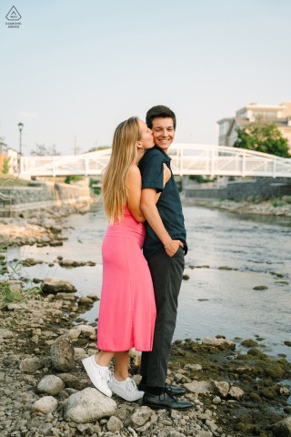 A couple stands together on a low water level of the river in Downtown West Bend, Wisconsin during an engagement photo shoot, with the picturesque bridge in the background