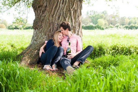 Una pareja disfrutando de una serena sesión de retratos en Fen Ditton Meadows, Cambridge, sentada junto a un majestuoso árbol mientras bebe champán en una pintoresca pradera