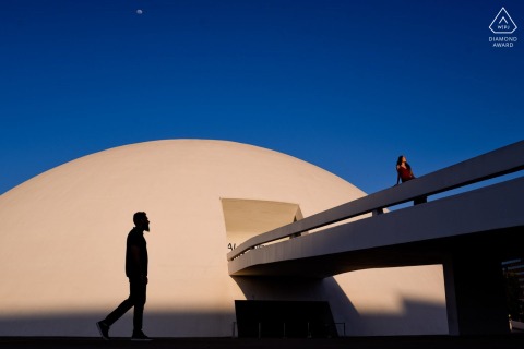 The photo captured in an engagement session at the National Museum in Brasilia. The image showcases the bride-to-be standing on a bridge against a beautiful deep blue sky, while her soon-to-be-husband walks below with a striking silhouette.