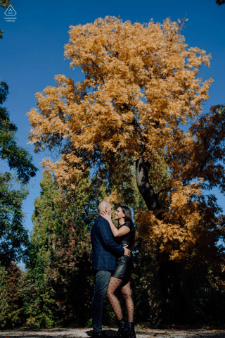 In the stunning Gödöllői kastély, a couple shares a sweet session as they embrace under a majestic tree adorned in vibrant, fall colors, captured by a Gödöllői kastély wedding photographer