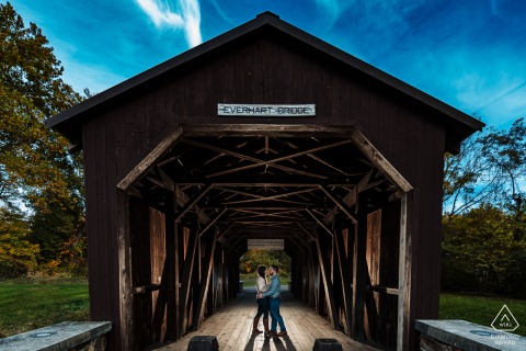 A couple about to get married in Harrisburg, PA, posed on a covered bridge in Fort Hunter for their engagement portrait session. The photographer used a remote strobe to create a backlit effect, adding to the impact of a symmetrical shot.