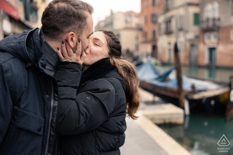 A beautiful pre-wedding engagement portrait captured in the picturesque Cannaregio district in Venice, Italy. The creative shot features a couple sharing a kiss, with the serene backdrop of canals and boats in the water behind them.